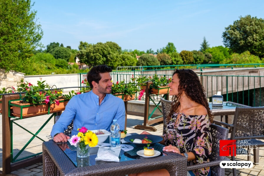 Coppia seduta al tavolo all'aperto, campo da golf sullo sfondo - logo scalapay e parc hotels italia